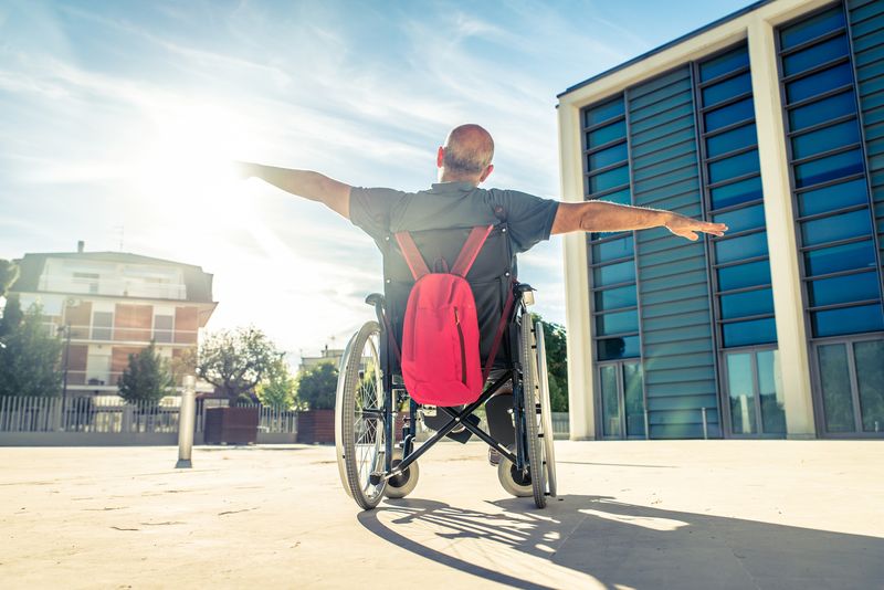 A man in a wheelchair throws his arms wide on a sunny plaza.