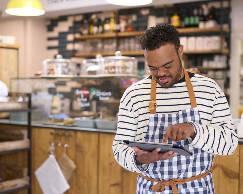 A man in a striped apron checks orders on a tablet in a cafe.