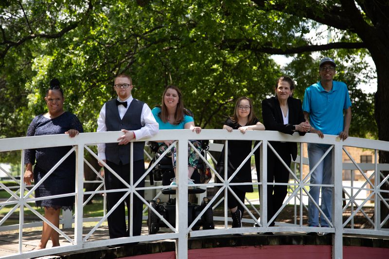 Friends lean on the railing of a park footbridge under shady trees.